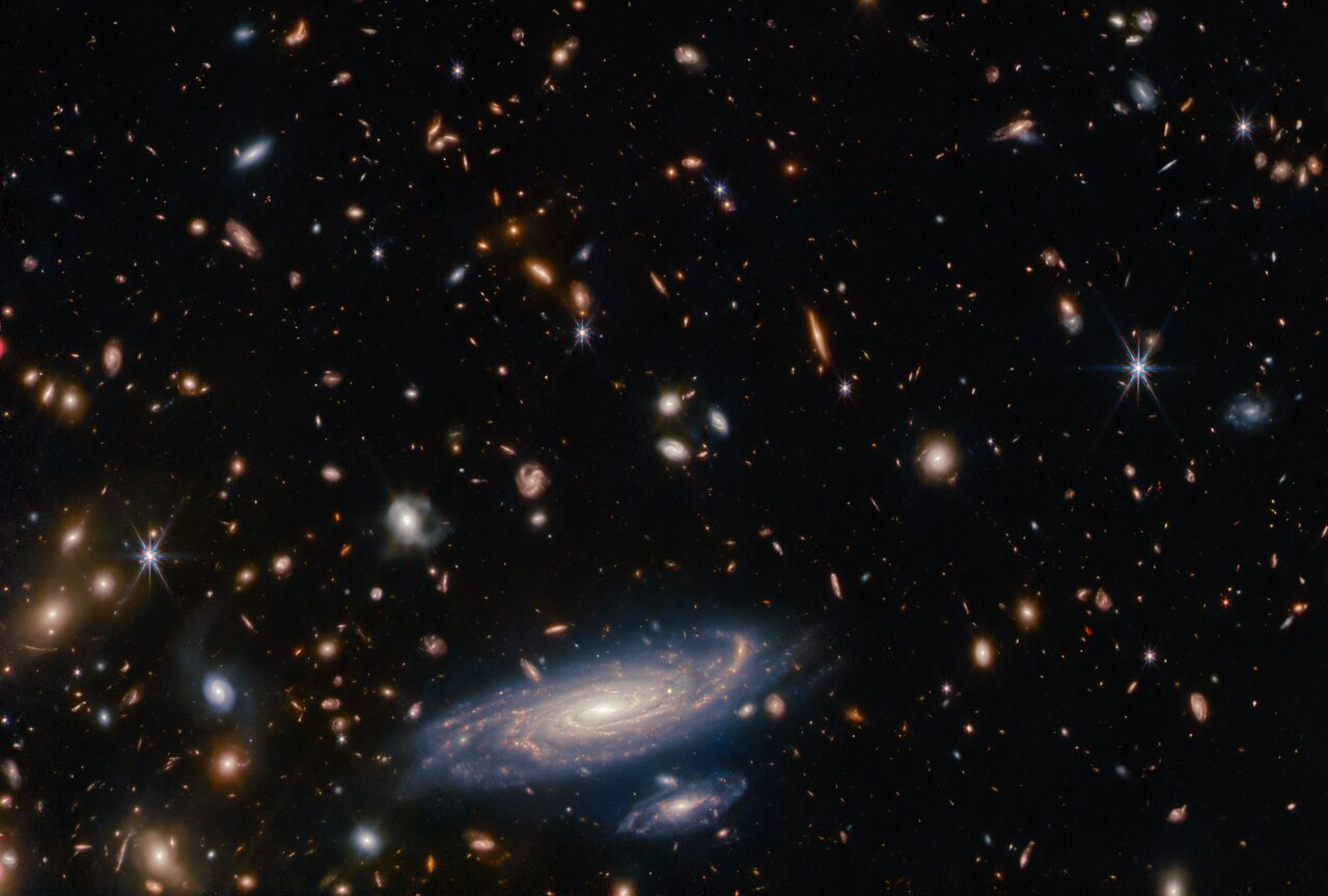 Many stars and galaxies lie on a dark background, in a variety of colours but mostly shades of orange. Some galaxies are large enough to make out spiral arms. Along the bottom of the frame is a large, detailed spiral galaxy seen at an oblique angle, with another galaxy about one-quarter the size just beneath it. Both have a brightly glowing core, and areas of star formation which light up their spiral arms.